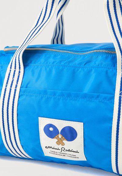Μπλε υφασμάτινη τσάντα duffel με λευκές και μπλε ριγωτές λαβές και μπροστινό ταμπελάκι που δείχνει δύο σταυρωτές μπλε ρακέτες επιτραπέζιας αντισφαίρισης.