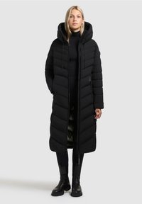 Parka nera imbottita con cappuccio, caratterizzata da un lungo design trapuntato e cuciture verticali. Indossata su un completo nero, abbinata a stivali neri.