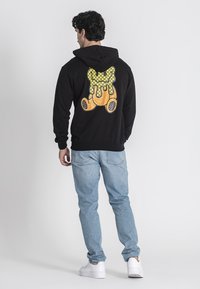 Felpa nera con un grafico colorato con un design giallo e marrone sulla schiena. Abbinata a jeans azzurri chiari e scarpe bianche.