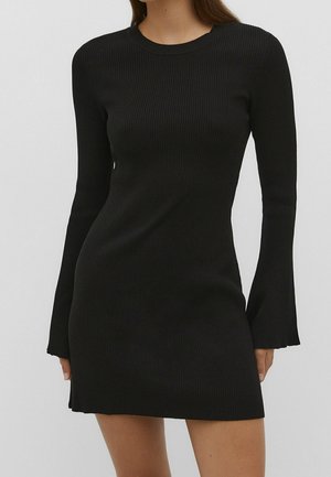 Femme portant une robe moulante en maille côtelée noire avec de longues manches évasées et un col rond sur un fond uni.