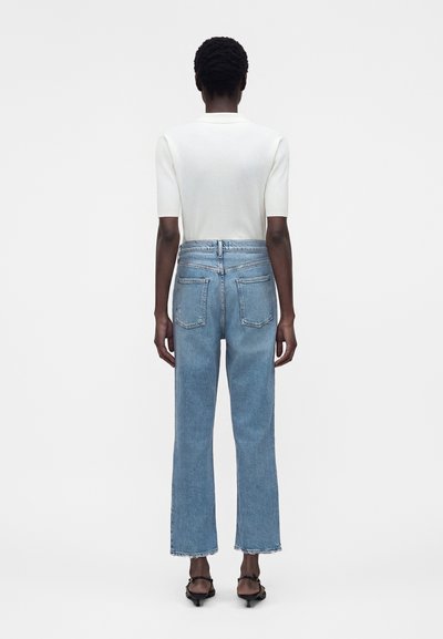 Άτομο που στέκεται με την πλάτη στην κάμερα, φορώντας λευκό ριπ κοντομάνικο τοπ, τζιν ψηλόμεσο και μαύρα σανδάλια σε λευκό φόντο.