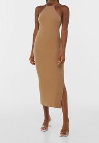Donna che indossa un vestito midi senza maniche, a coste, color sabbia, con scollatura a halter e uno spacco laterale, abbinato a sandali con tacco a spillo trasparenti.