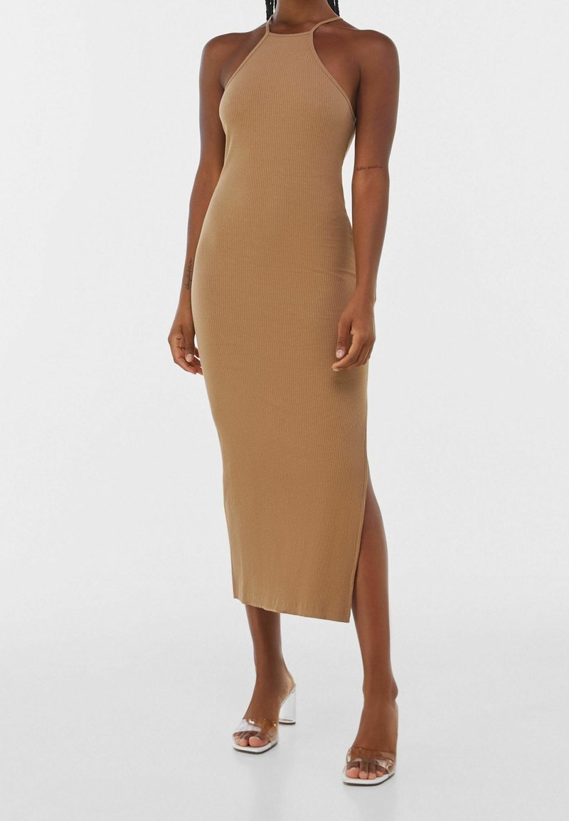 Donna che indossa un vestito midi senza maniche, a coste, color sabbia, con scollatura a halter e uno spacco laterale, abbinato a sandali con tacco a spillo trasparenti.