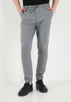 Person wearing slim-fit grey trousers with a drawstring and button waist, black sneakers, and hands in pockets against a plain background.