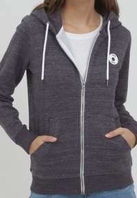 Mujer con una sudadera con cremallera gris oscuro y cordones blancos, y una camiseta blanca, con las manos en los bolsillos delanteros, visible desde el cuello hasta la cadera.