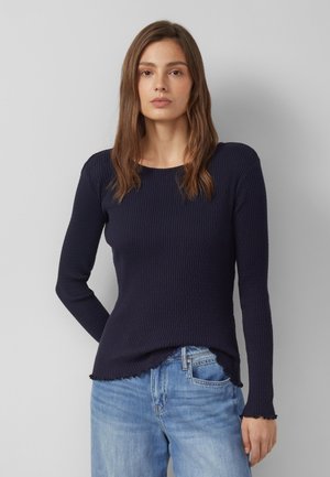 Vrouw met lang bruin haar, gekleed in een donkerblauw geribd shirt met lange mouwen en lichtblauwe spijkerbroek, staand tegen een effen lichtgrijze achtergrond.