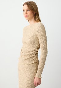 Top beige strutturato con maniche lunghe, presenta un design arricciato sul lato, realizzato in un tessuto elasticizzato, abbinato a una gonna coordinata.