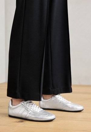 Metallic silver sneakers with a smooth texture, featuring white laces and side stripes, paired with wide black pants with a cropped hem.