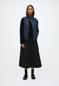 Ναυτικό κομμένο γιλέκο με ψηλό γιακά και τσέπη σε σμαραγδί απόχρωση, φορεμένο πάνω από ένα μαύρο μπλουζάκι με μακριά μανίκια και μαύρη μακρύ φούστα, σε συνδυασμό με μαύρα παπούτσια.