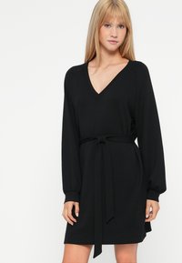 Vestido negro con escote en V, mangas largas y lazo en la cintura. Hecho de una tela suave, con un corte relajado y puños acanalados.