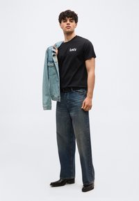 Νέος άνδρας που φοράει μαύρο t-shirt Levi's, μπλε τζιν, μαύρες μπότες, κρατώντας ένα ανοιχτό μπλε τζιν μπουφάν πάνω σε έναν ώμο.