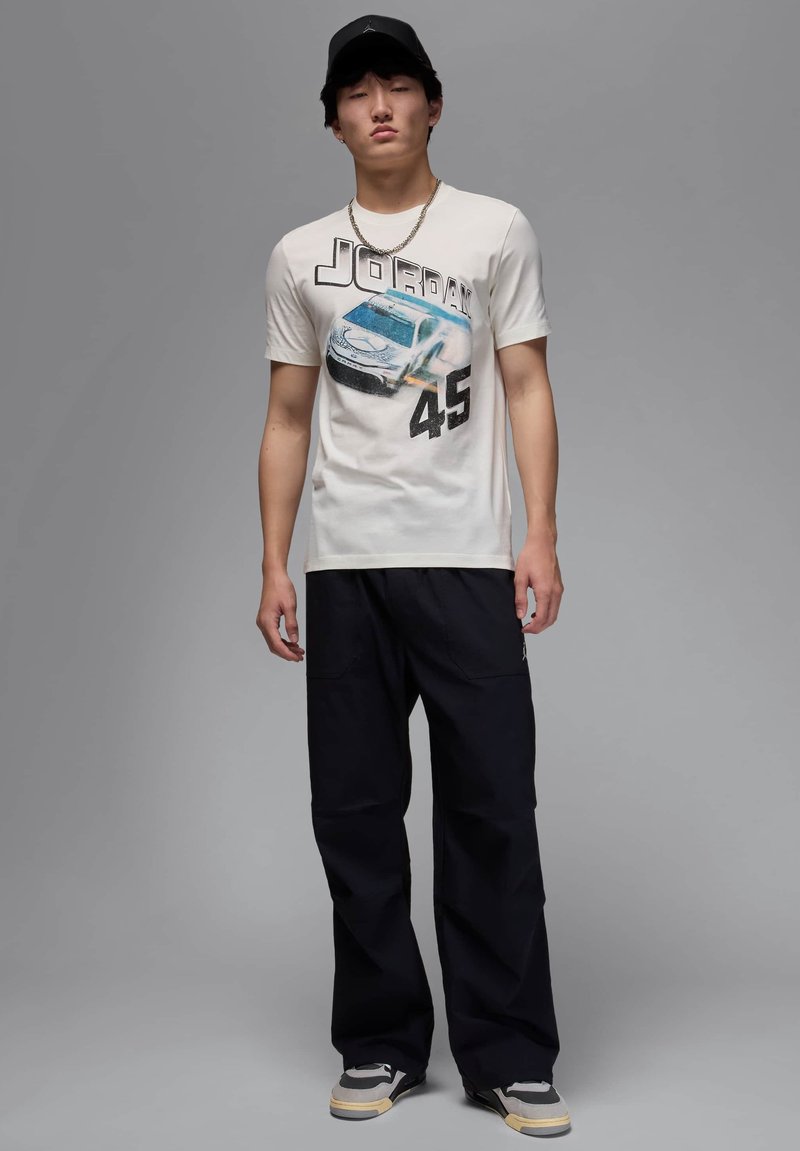 Maglietta grafica bianca con un design di un'auto e il testo "JORDAN 45". Indossata con pantaloni neri larghi e sneakers grigie con accenti neri.