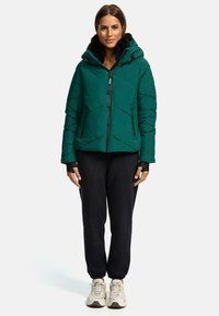 Green puffer jacket with a zip front, hood, and contrasting black cuffs. Paired with black joggers and light-colored sneakers.