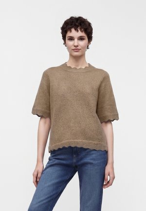 Ung person med kort krøllet hår iført en brun tekstureret kortærmet sweater og blå jeans, stående mod en ensfarvet baggrund.
