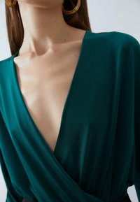 Tee-farbene Wickelbluse mit tiefem V-Ausschnitt, aus glattem Stoff gefertigt. Ein dunkler schwarzer Taillengürtel betont die Silhouette; das Model trägt große goldene Creolen.