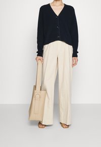 Persona che indossa un maglione nero con bottoni, pantaloni larghi beige, tiene una borsa tote beige, in piedi su un pavimento grigio contro un muro bianco.