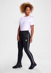 Zwarte trainingsbroek met elastische tailleband, met een grijs chevronpatroon langs de zijkanten. Gecombineerd met een witte T-shirt met ronde hals.