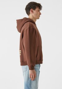 Ruda kapucha sukurta iš minkštos medžiagos, laisvu siluetu. Savybės: priekinė kišenė, suveržti rankogaliai ir spaudinys šone.