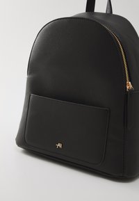 Sac à dos noir de forme arrondie, fabriqué en matériau texturé, doté d'une fermeture éclair dorée et d'une poche avant avec un discret logo.