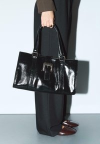 Sac à main en cuir verni noir avec deux poignées, de forme rectangulaire, présentant un détail de boucle carrée en argent, associé à un pantalon noir et des chaussures marron.