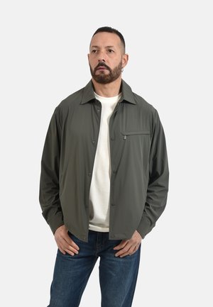 Uomo con barba che indossa una giacca verde scuro con bottoni sopra una camicia bianca e jeans blu, in piedi con le mani sui fianchi davanti a uno sfondo uniforme.