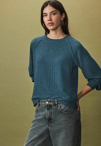 Teal gemêleerd sweatshirt met raglanmouwen, gecombineerd met hoge taille denim jeans, tegen een gedempte groene achtergrond.