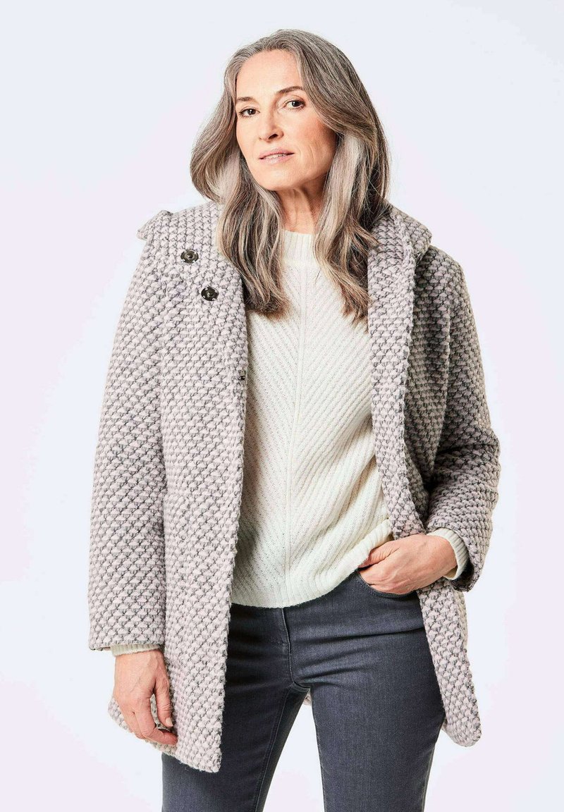 Manteau en laine grise texturée avec un tissage à motifs, grand col et deux fermetures à bouton, associé à un pull léger en tricot et un jean foncé.