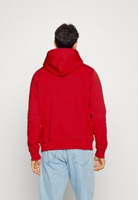 Sweat à capuche rouge avec une grande capuche et des poignets côtelés. Le tissu semble doux et texturé, avec un cordon de serrage et une poche kangourou non visible.