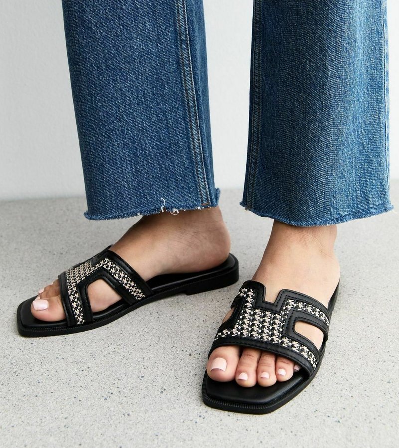 Svarta slip-on sandaler med houndstooth-mönster framtill, fyrkantig tåsform, på en bar fot bredvid fransade blå denimbyxor.