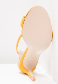 Sandale à talon haut en suede jaune, présentant un talon fin, un dos ouvert et une sangle ajustable. La semelle lisse de couleur claire est visible.