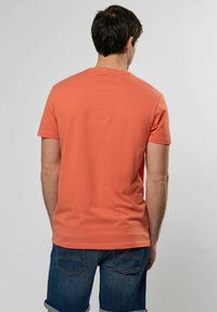 Krátký rukáv tričko z korálové bavlny, jednobarevný design, kulatý výstřih, hladká textura. Nosené s denimovými šortkami, zadní pohled na světlem pozadí.