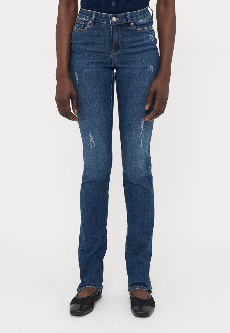 Oseba, oblečena v oprijete modre kavbojke s svetlimi poškodovanji, v kombinaciji s črnimi ravnimi čevlji, stoji ob pusti beli ozadju.