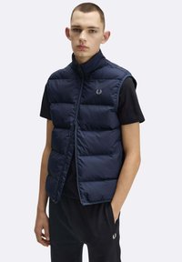 Smanicato trapuntato imbottito blu navy, design senza maniche, colletto alto, cerniera frontale, con logo marchiato sopra il petto sinistro. Indossato sopra una t-shirt nera.