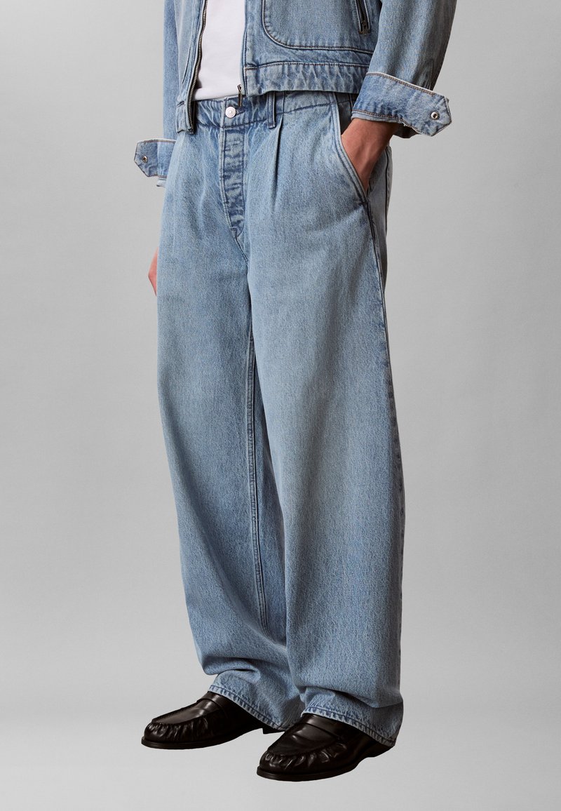 Άτομο που φοράει ανοιχτό μπλε τζιν παντελόνι με φαρδιά μπατζάκια, τζιν μπουφάν, λευκό πουκάμισο, μαύρα μοκασίνια, στέκεται με τα χέρια στις τσέπες μπροστά σε γκρι φόντο.