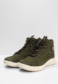 Zapatillas altas de color verde oliva, hechas de material sintético con patrones texturizados, con acentos en negro, suelas texturizadas y etiquetas de marca.