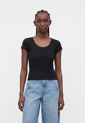 Femme aux cheveux tressés portant un t-shirt noir ajusté à manches courtes et un jean taille haute bleu clair, debout devant un fond blanc uni.