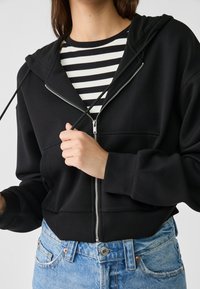 Sudadera negra con cremallera y capucha, con cremallera plateada y bolsillos tipo canguro, llevada sobre una camiseta de rayas blancas y negras, combinada con vaqueros azules de denim.