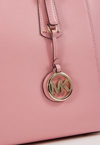 Gros plan d’un sac à main en cuir rose texturé avec un pendentif circulaire doré du logo Michael Kors suspendu à une sangle cousue.