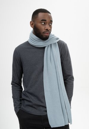 Jonge man met baard, gekleed in een donkergrijs shirt met lange mouwen en een lichtblauwe gebreide sjaal, kijkt opzij met zijn handen in zijn zakken.