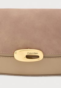 Calvin Kleini käekott, mille pealmine osa on pehmest tuhmunud pruunist seemisnahast, alus silest beežist nahast ning kullast ovaalse detailiga, millel on logo.