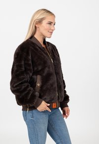 Bruine nepbont bomberjack met ribgebreide manchetten en zoom, gouden rits, zijzakken en oranje labeldetail. Draagt met blauwe jeans.