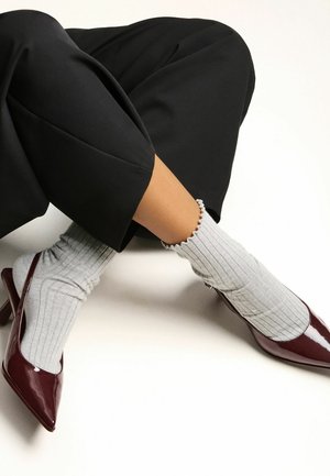 Personne portant des chaussettes gris côtelées avec des bords froncés, associées à des escarpins en cuir verni bordeaux et à un pantalon noir coupé, sur fond blanc.