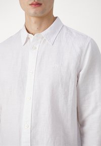 Camicia button-up bianca in lino con una sottile trama, dotata di colletto standard e logo del marchio ricamato sul petto.
