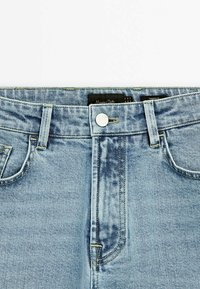 Gros plan de la ceinture d'un jean ajusté bleu clair, avec fermeture par bouton et passants de ceinture.