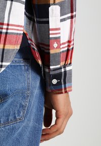 Camicia a maniche lunghe a scacchi in bianco, blu navy, rosso e giallo. Tessuto in cotone con polsini abbottonati e colletto classico. Indossata con jeans in denim blu.