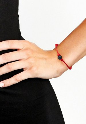 Bracelet élastique rouge avec une perle noire centrale, porté sur une main contre un fond noir. Design simple avec un ajustement réglable.