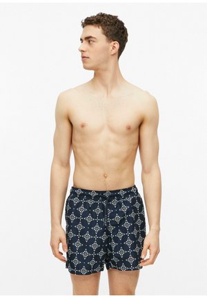 Jeune homme aux cheveux courts et bouclés portant un short de bain bleu marine avec des motifs géométriques blancs, regardant vers la gauche sur un fond blanc uni.