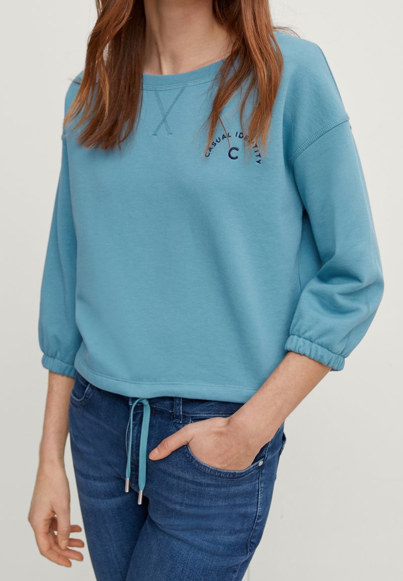 Sweat-shirt bleu clair à coupe décontractée, manches trois-quarts, ourlet avec cordon de serrage et texte brodé. Texture de tissu douce et lisse.