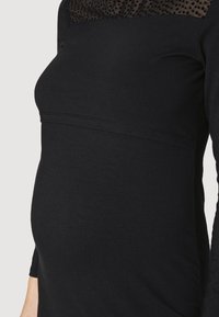 Musta äitiysmekko, jossa on tiukka bodice, teksturoitu verkko-osa, jossa on pilkkuja, ja pitkät hihat. Silkkinen kangas, jossa on venyvyyttä.