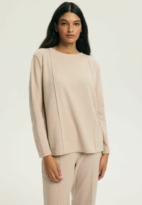 Beżowa bluza z długim rękawem wykonana z miękkiego materiału, z zaokrąglonym dekoltem i pionowymi przeszyciami. Dobrana do niej beżowe spodnie w tym samym odcieniu.